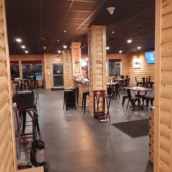 Interior view of a restaurant with wooden walls, dark tables, chairs, and a bar area.